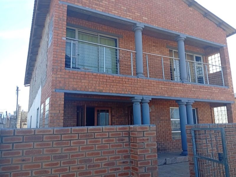 A double-story face brick house featuring a front balcony with concrete columns, stainless steel balustrades, and a secure gated entrance.