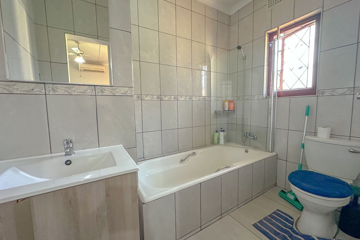 A functional bathroom featuring a built-in bathtub with shower screen, vanity basin, and toilet, finished with neutral wall and floor tiling.