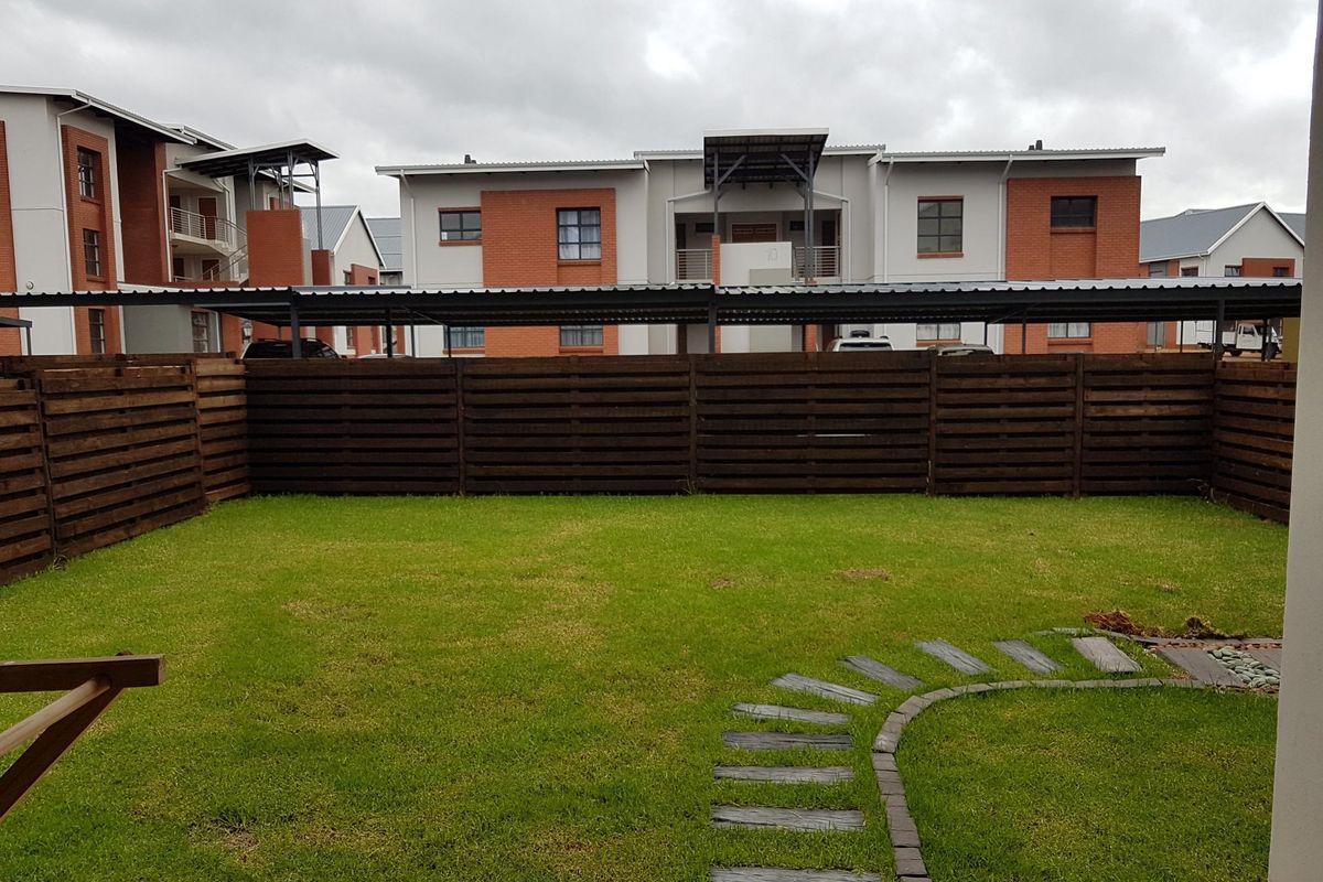 A private rear garden featuring a well-maintained lawn, stone paver pathway, and timber slat fencing, set against the backdrop of the townhouse complex.