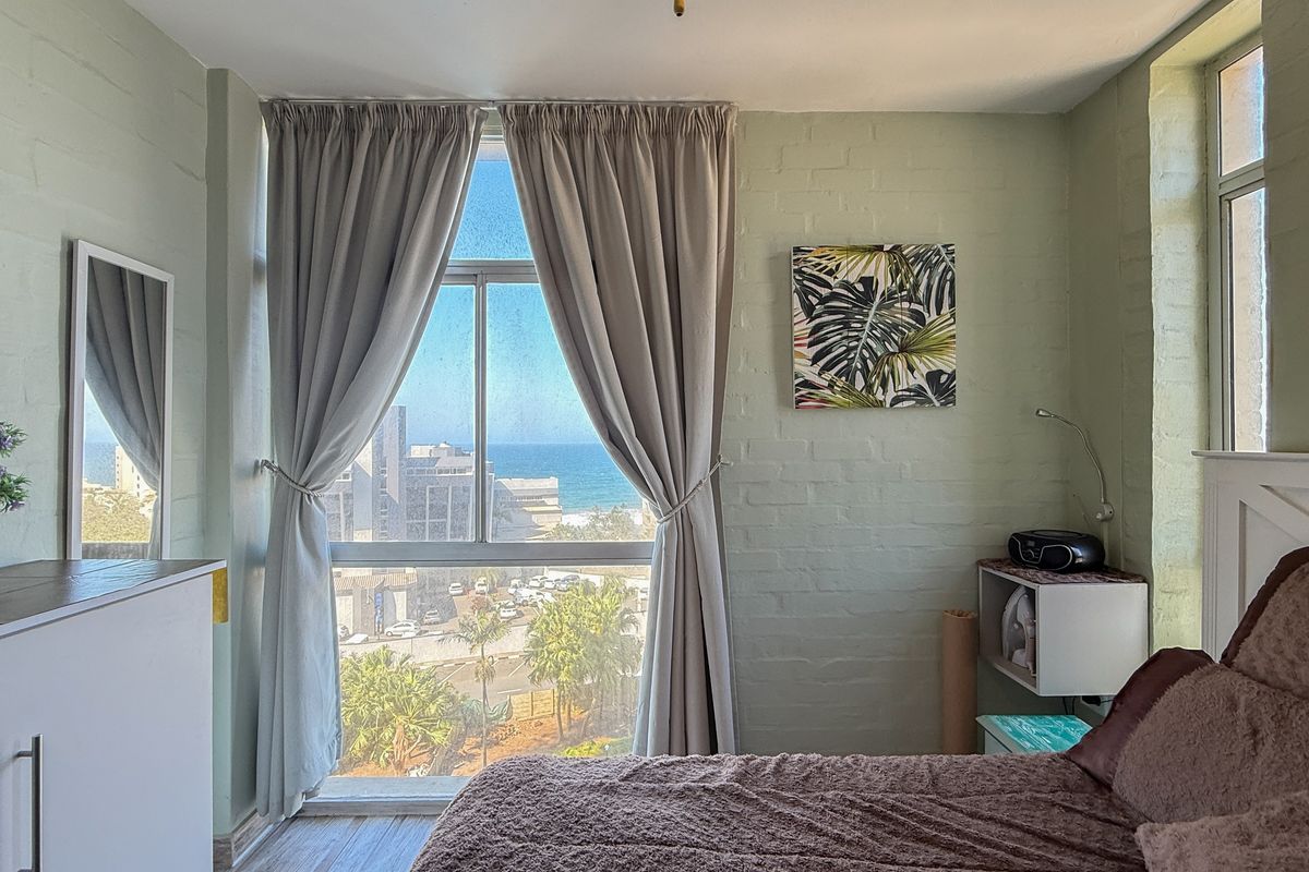 A bedroom featuring painted brick walls, wood-look flooring, and a large window offering a scenic coastal and urban view.
