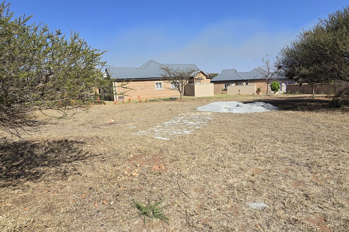 A view of a vacant residential plot featuring dry grass, mature trees, and a small pile of construction materials.