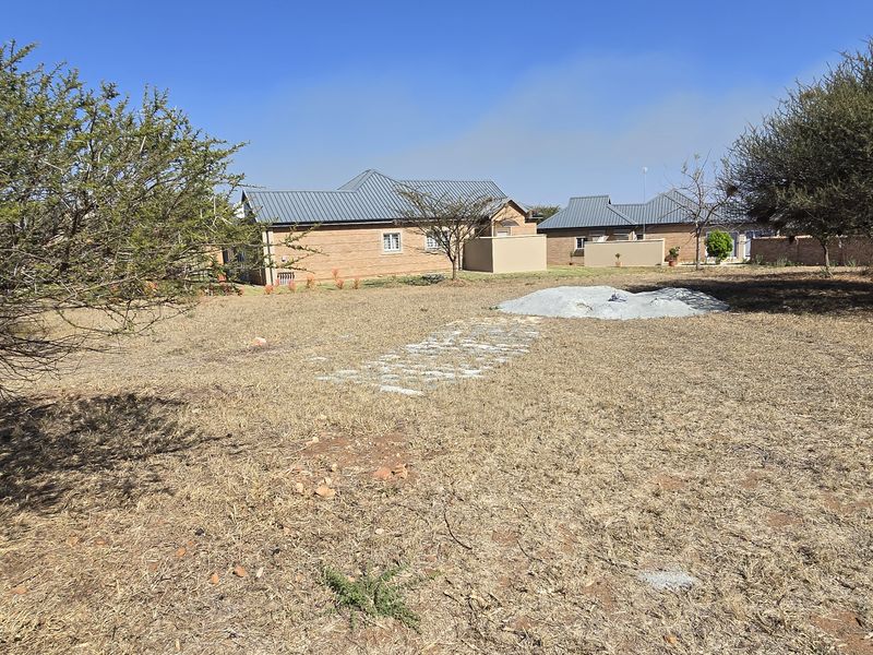 A view of a vacant residential plot featuring dry grass, mature trees, and a small pile of construction materials.