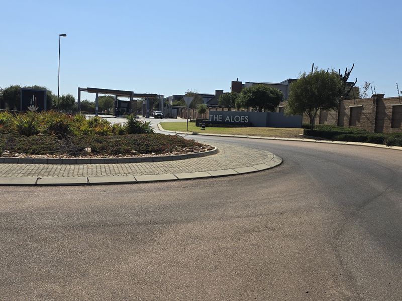 View of the entrance and security gatehouse for The Aloes residential estate, featuring paved roads and landscaped roundabout.