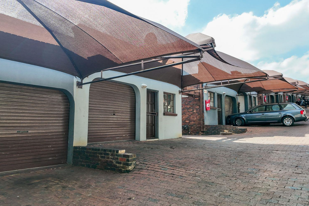 A row of apartment garages featuring roller shutter doors and overhead shade cloth structures, set within a paved residential parking area.