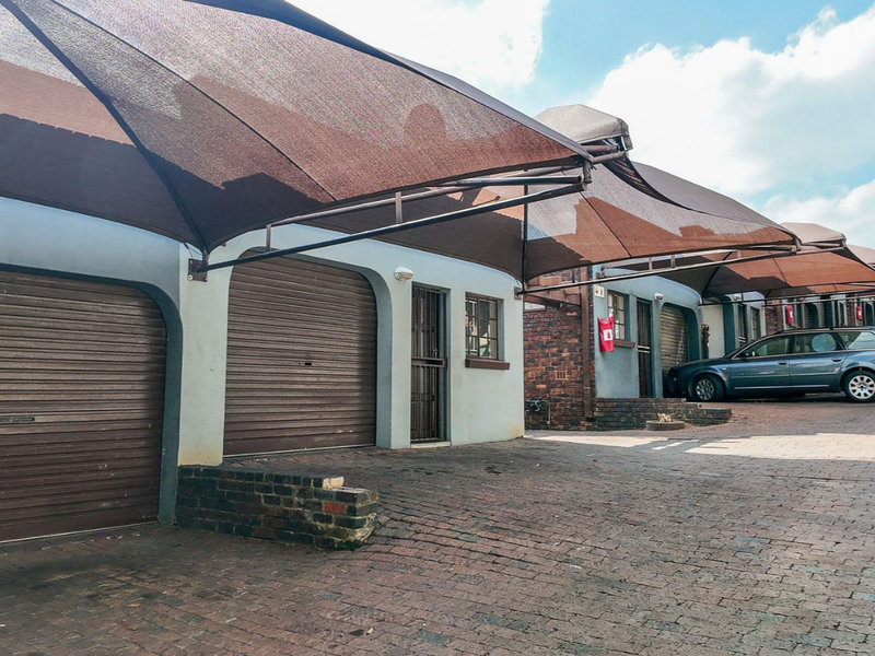 A row of apartment garages featuring roller shutter doors and overhead shade cloth structures, set within a paved residential parking area.