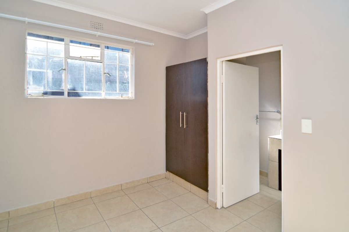 A bright, empty bedroom featuring tiled flooring, a built-in dark wood wardrobe, and an adjoining bathroom entrance.