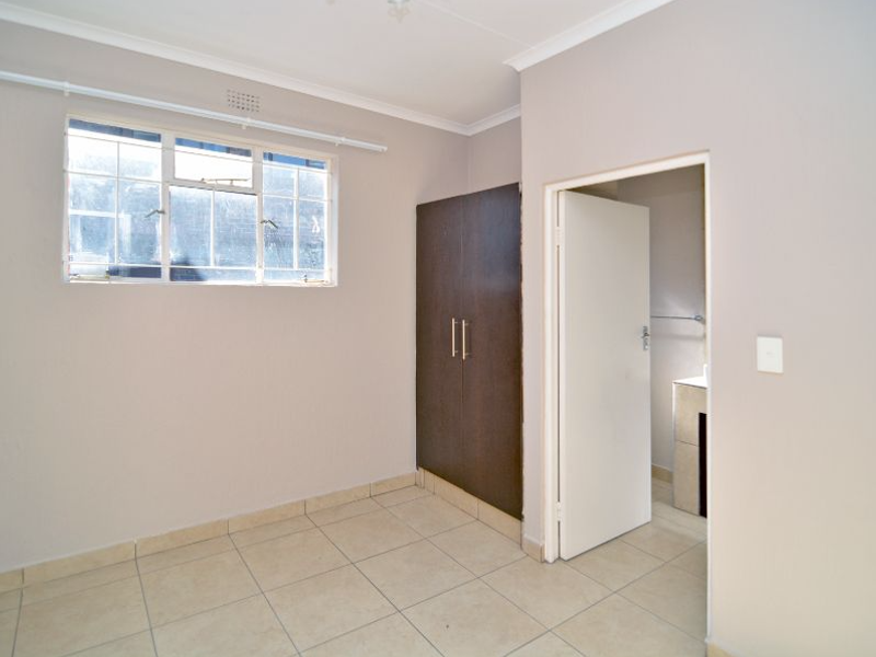 A bright, empty bedroom featuring tiled flooring, a built-in dark wood wardrobe, and an adjoining bathroom entrance.
