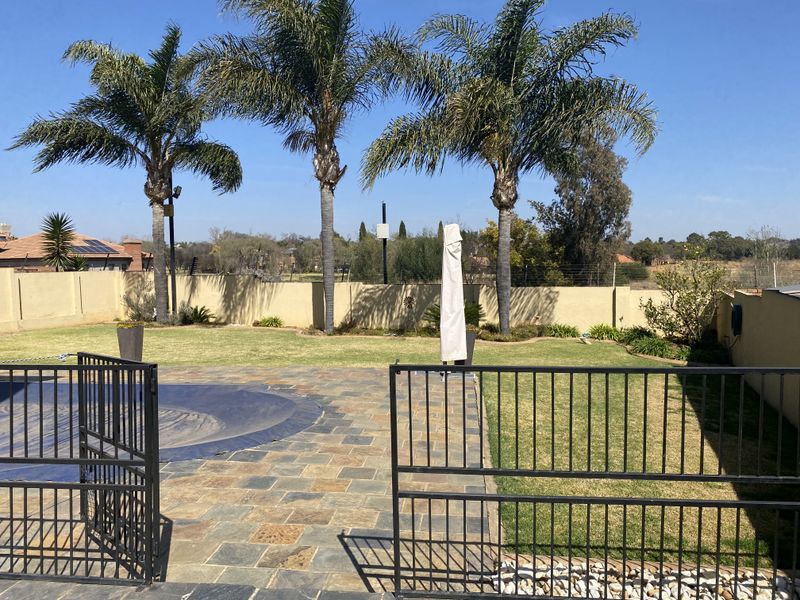 A well-maintained outdoor pool area featuring stone paving, perimeter walling, metal security fencing, and established palm trees in a sunny garden setting.