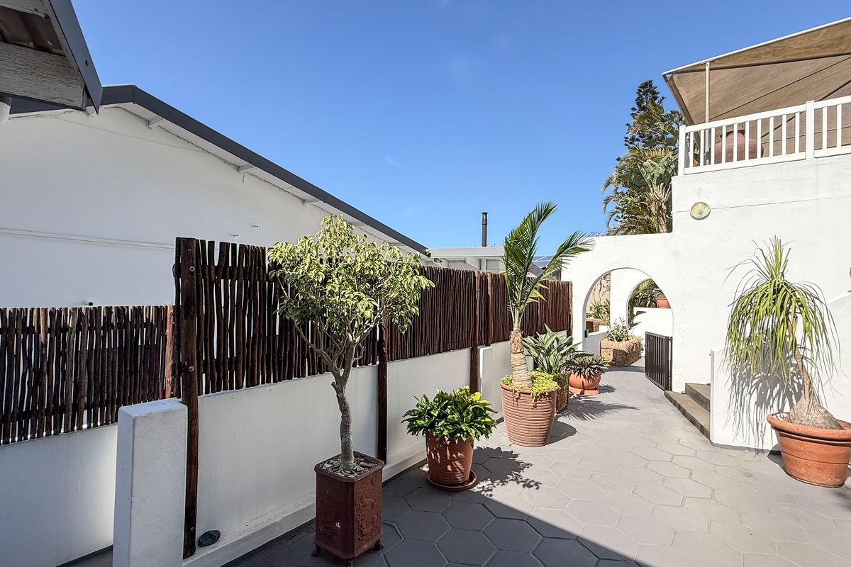 A paved outdoor walkway featuring white rendered walls, a rustic timber screen fence, and architectural archways leading to a rear garden area.