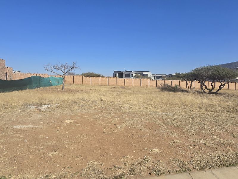 A vacant residential plot featuring dry grass, perimeter boundary walls, and established trees, situated in a developed neighborhood.