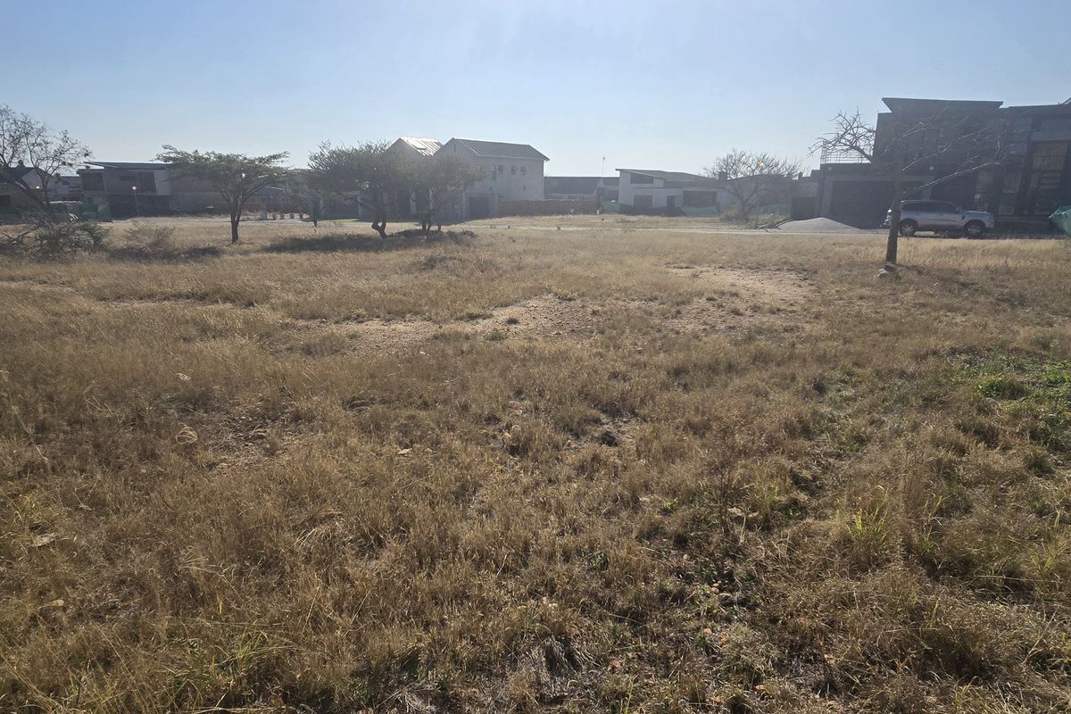 A vacant residential plot of land featuring dry grass, scattered trees, and surrounding residential development in a suburban estate setting.