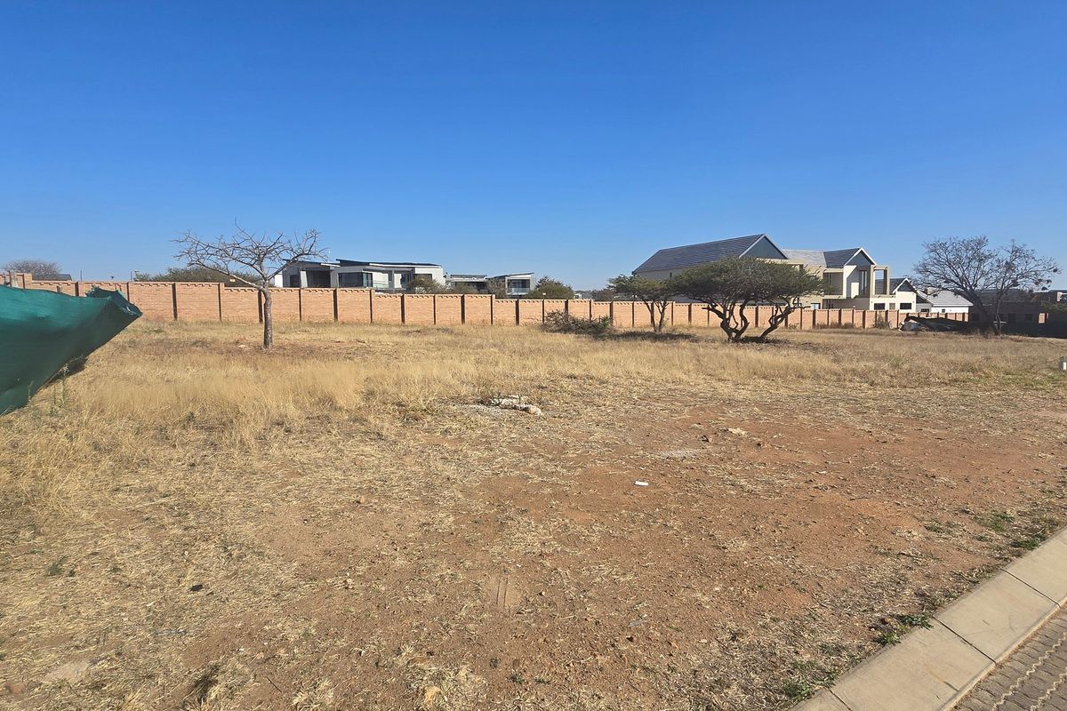 A vacant residential plot featuring dry grass, mature trees, and a perimeter brick wall, situated adjacent to a paved street.