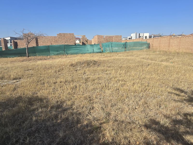 A vacant residential plot featuring dry grass, perimeter brick walling, and temporary green construction screening.