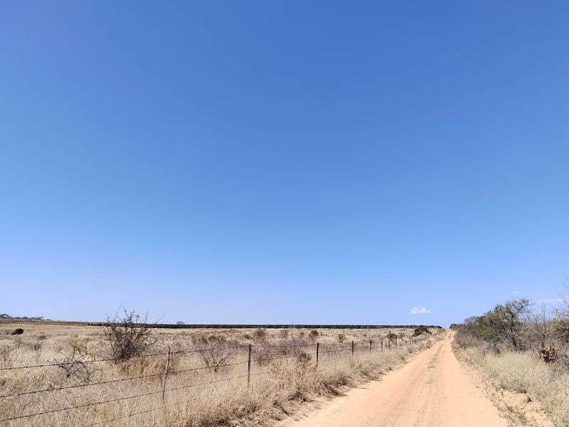 A view of a dirt access road running alongside a wire fence on a rural farm property under a clear blue sky.