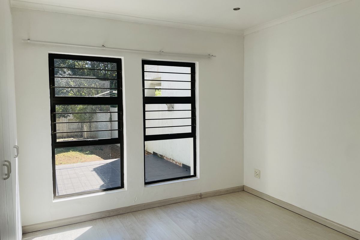 A bright, empty bedroom featuring light-toned laminate flooring, white walls, and black-framed windows with integrated security bars.