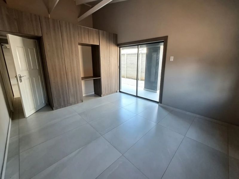 A bright, empty bedroom featuring modern tiled flooring, extensive built-in cabinetry with a desk nook, and a sliding door leading to an outdoor area.