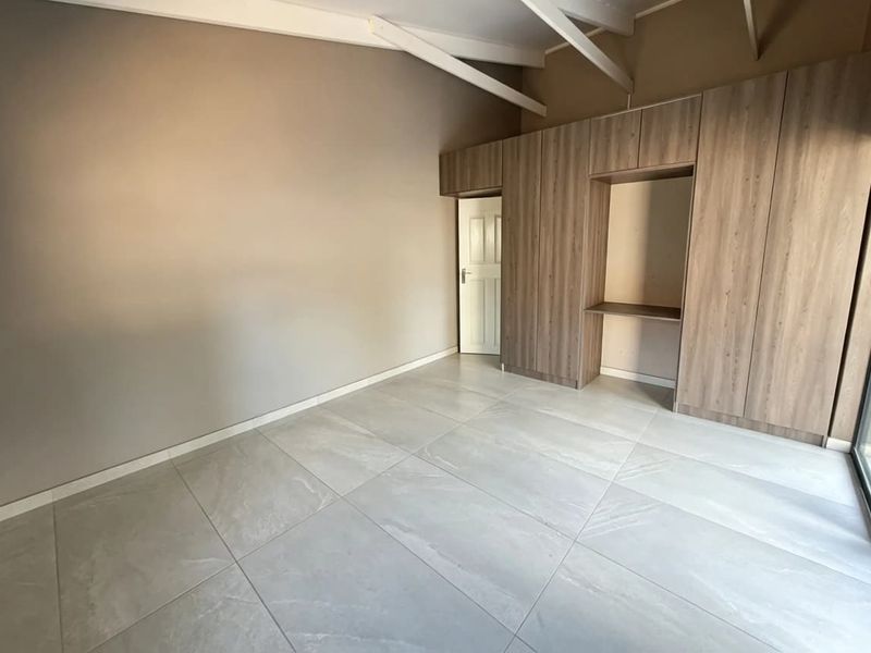 A spacious, empty bedroom featuring modern tiled flooring, built-in wooden cupboards, and exposed ceiling rafters, offering a clean and neutral living space.