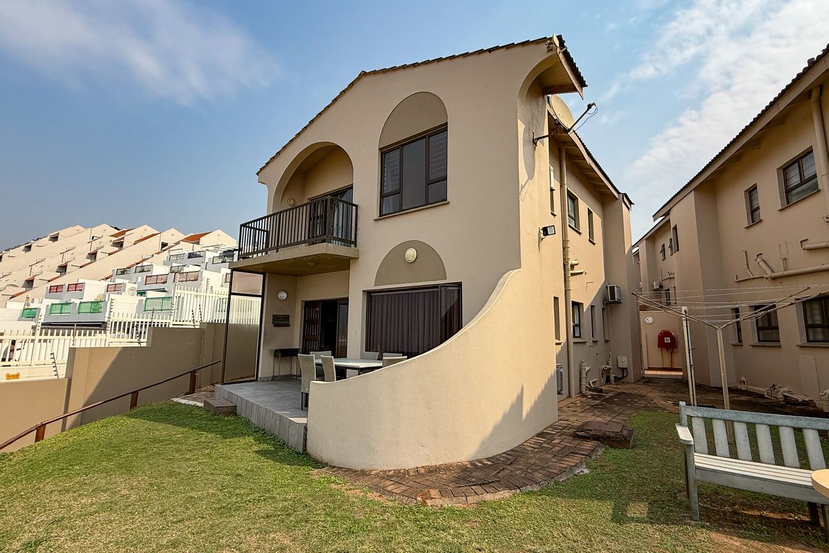 Rear view of a multi-story apartment unit featuring a private patio, balcony, and small garden area within a residential complex.