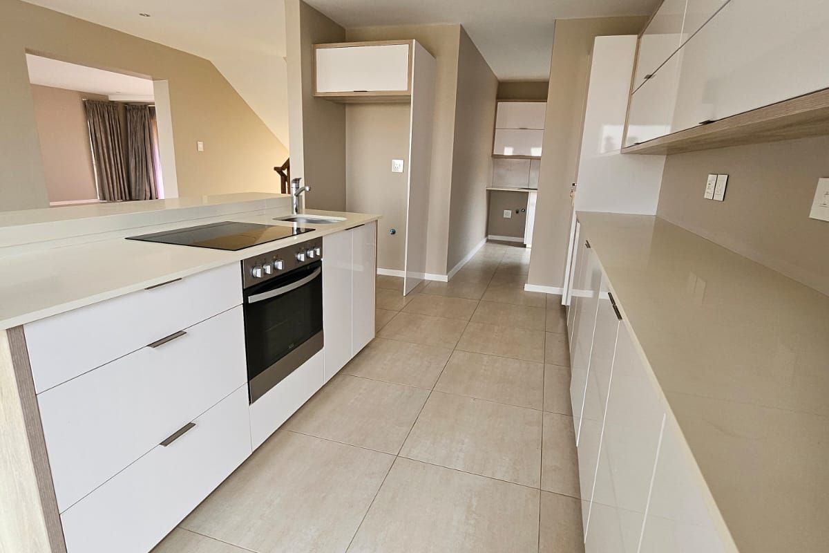 Modern kitchen featuring sleek white cabinetry, stone countertops, built-in oven, electric hob, and neutral tiled flooring in an open-plan layout.