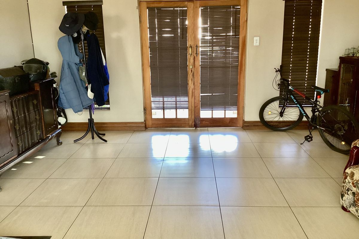 A living area featuring large ceramic floor tiles, wooden double doors leading outside, and fitted wooden blinds on the windows.