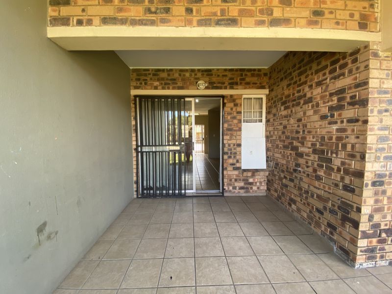 A covered patio area featuring tiled flooring, exposed brick walls, and a secure sliding glass door entrance into the home.