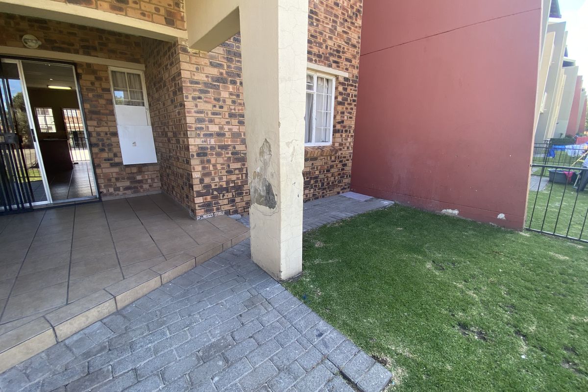 Rear exterior view of a townhouse featuring a tiled patio, paved walkway, small lawn, and brick facade with visible maintenance required on the structural pillar.