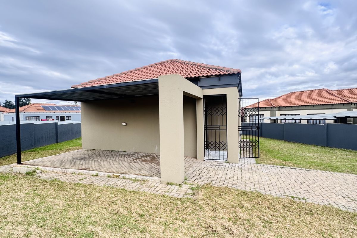 A rear view of the property featuring a covered patio area with paved flooring, a security gate, and a surrounding garden with boundary walls.