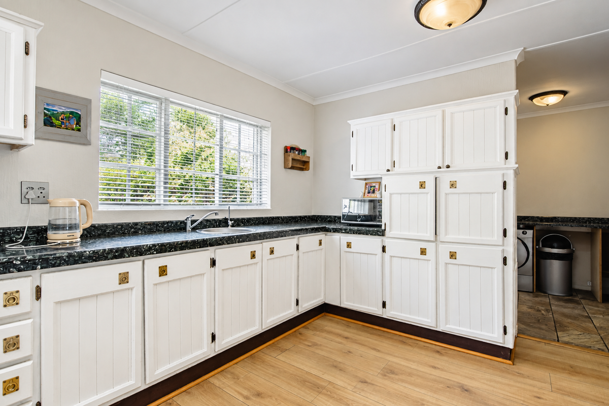 A functional kitchen featuring white cabinetry, dark laminate countertops, a stainless steel sink, and light wood-look laminate flooring with natural light from the window.