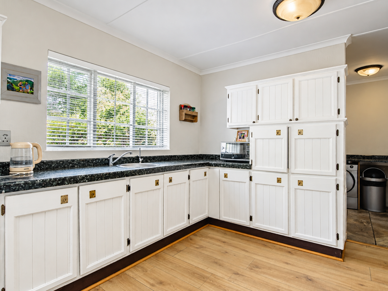 A functional kitchen featuring white cabinetry, dark laminate countertops, a stainless steel sink, and light wood-look laminate flooring with natural light from the window.