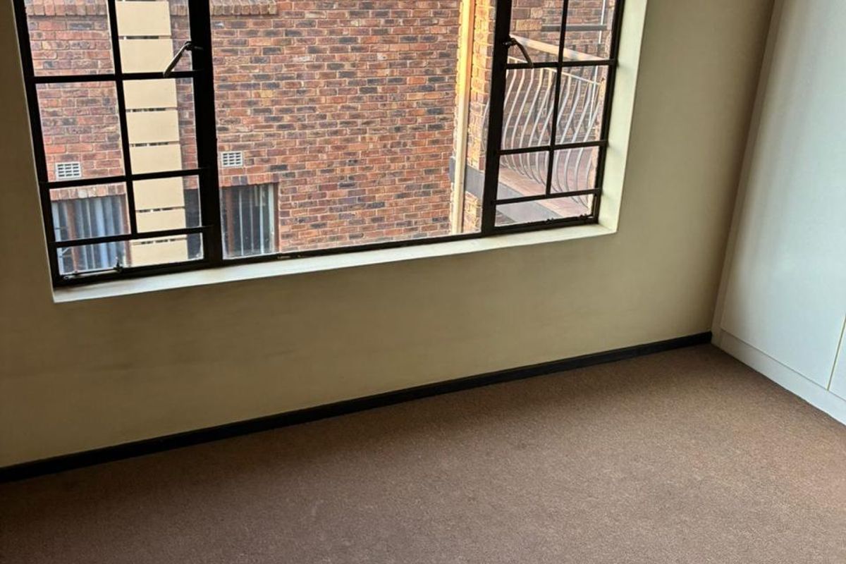 A vacant bedroom featuring brown carpeting, built-in cupboards, and large windows overlooking the adjacent building exterior.