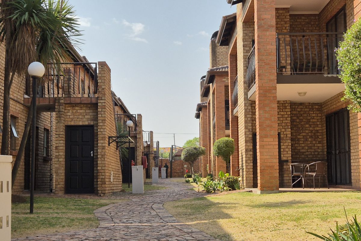 A view of the townhouse complex exterior featuring face brick facades, paved walkways, and private balconies overlooking a landscaped garden area.