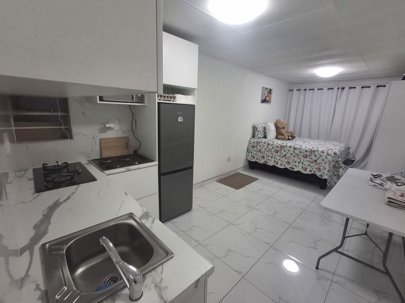 Open-plan studio apartment featuring a compact kitchen with marble-look countertops, dual hobs, and tiled flooring throughout the living and sleeping area.