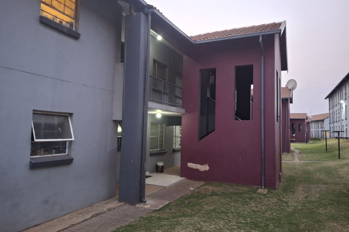 Exterior view of an apartment building complex featuring painted masonry walls, tiled roofing, and paved walkways with visible maintenance required on the exterior plaster.