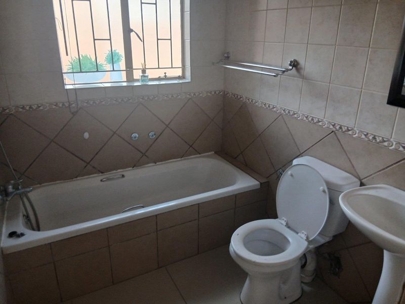 A functional bathroom featuring a built-in bathtub, pedestal basin, and toilet, finished with neutral ceramic wall tiles and security bars on the window.