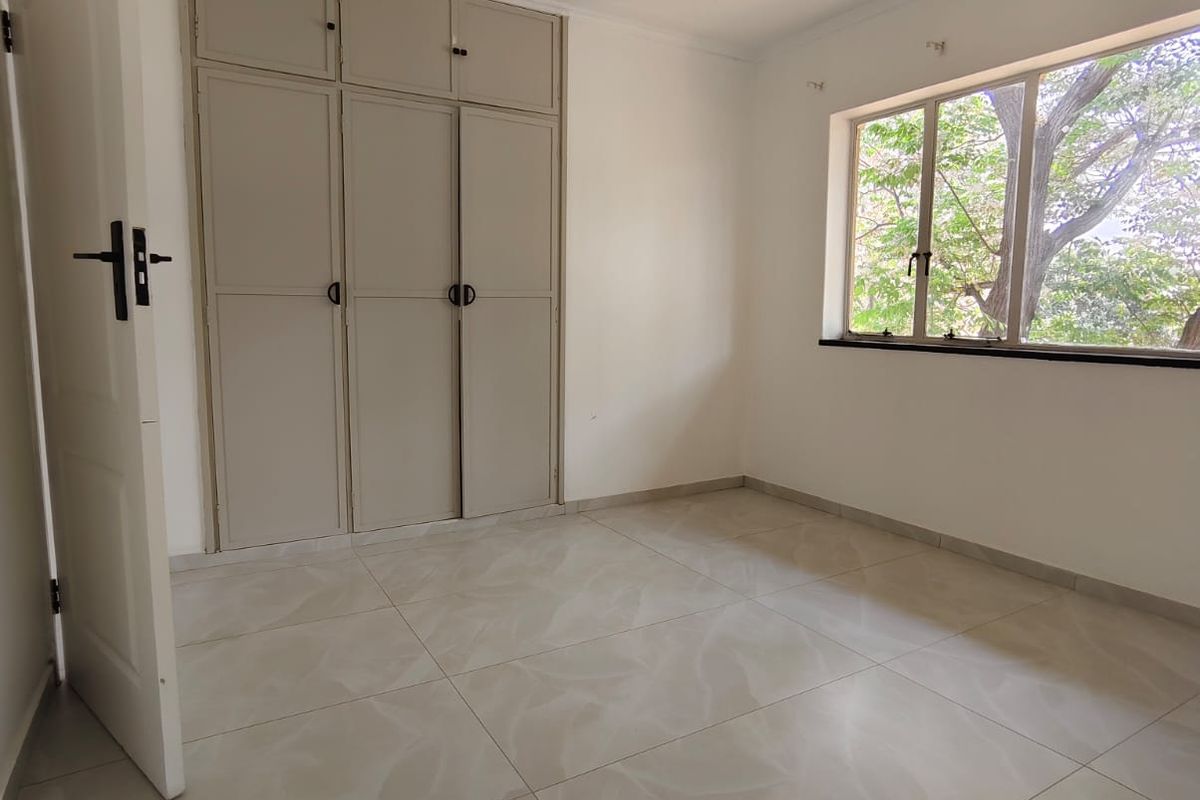 A bright, empty bedroom featuring neutral floor tiles, built-in cupboards, and a large window providing natural light and garden views.