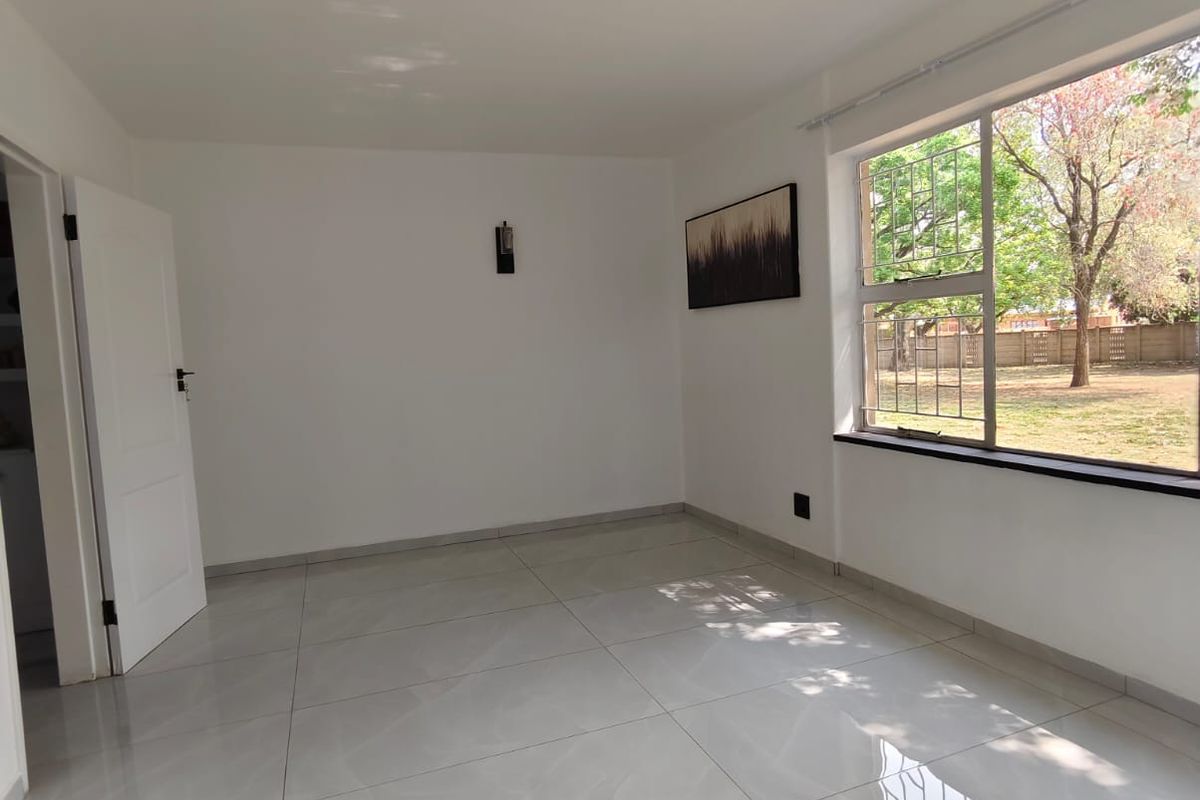 A bright, empty living room featuring large format polished porcelain floor tiles, a large window with security bars, and neutral white walls.