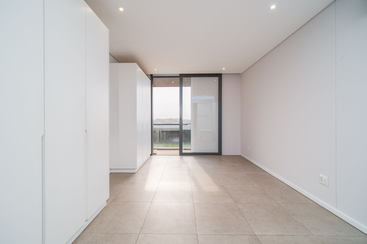 A bright, modern, and empty bedroom featuring neutral tiled flooring, built-in cupboards, and a sliding glass door leading to an outdoor area.