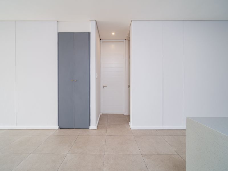 A clean, minimalist hallway featuring neutral tiled flooring, a grey built-in cupboard, and a white internal door with recessed lighting.