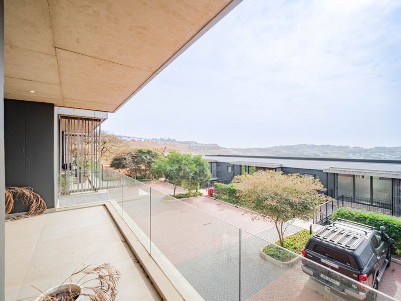 A modern, spacious balcony featuring a sleek glass balustrade, tiled flooring, and a concrete ceiling, offering an elevated view of the complex grounds.