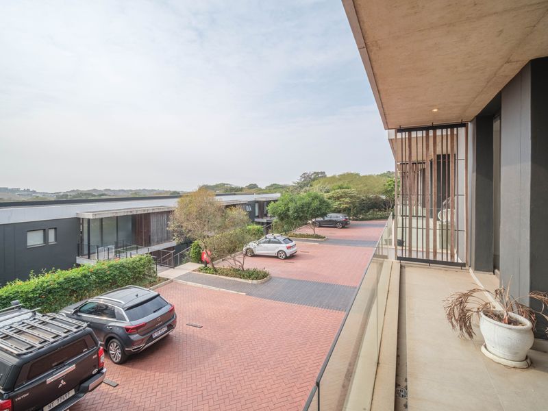 A modern, covered balcony featuring a glass balustrade and concrete ceiling, overlooking a paved parking area and landscaped surroundings.