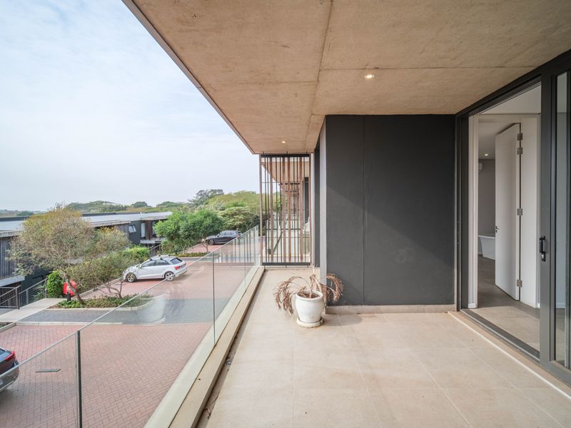 A modern, covered balcony featuring a glass balustrade, tiled flooring, and recessed lighting, offering an elevated view of the surrounding complex.