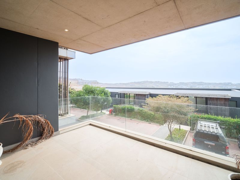 A spacious, modern balcony featuring a sleek glass balustrade, tiled flooring, and a concrete ceiling, offering an open view of the surrounding estate.
