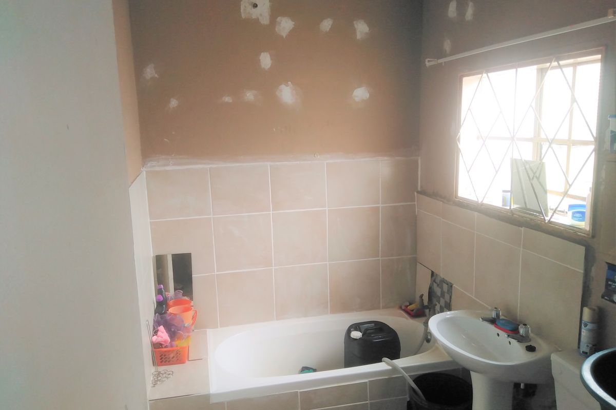 A bathroom in need of renovation, featuring a bathtub, pedestal basin, and window with security bars, currently showing unfinished wall repairs.