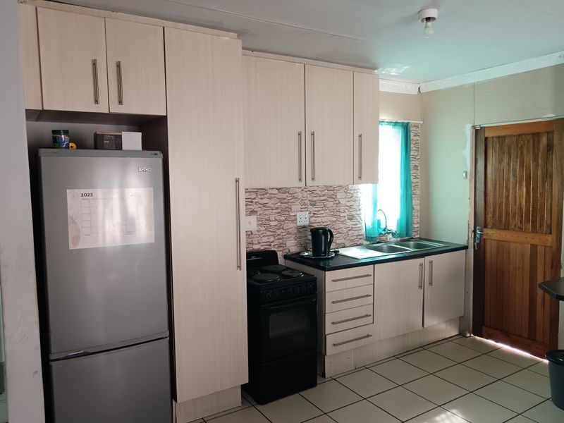 A functional kitchen featuring light-toned cabinetry, a dark countertop, a built-in sink, and a freestanding stove, with direct access to an exterior wooden door.