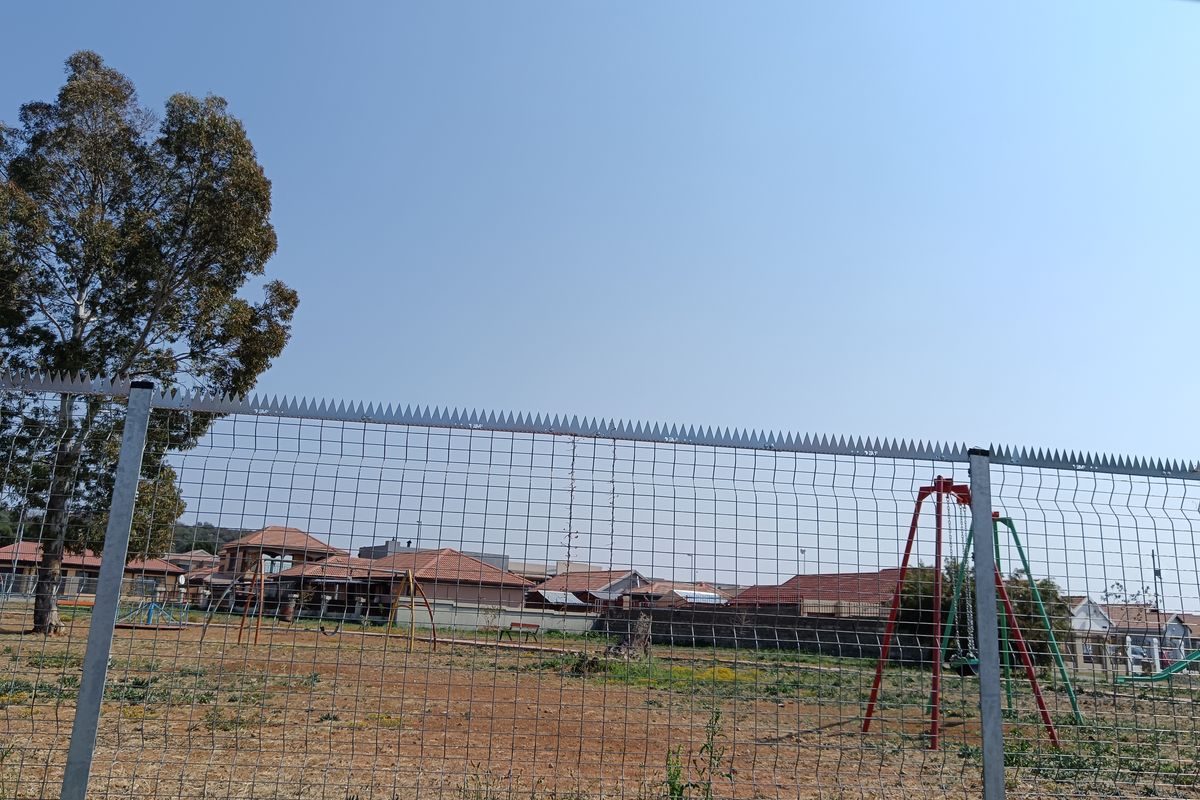 View of a communal outdoor area featuring a wire mesh security fence with spiked topping and a playground area in the background.