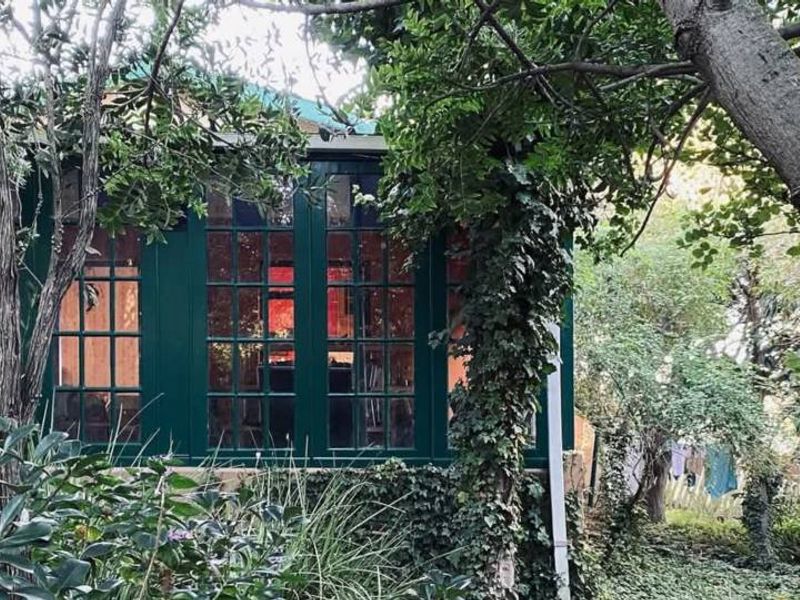 A view of the rear exterior of the property, featuring dark-framed glass windows and doors surrounded by mature trees and garden foliage.