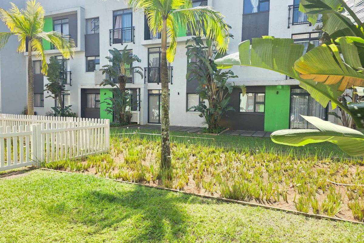 A view of the apartment complex exterior featuring a landscaped garden area, white picket fencing, and modern building facade with balconies.