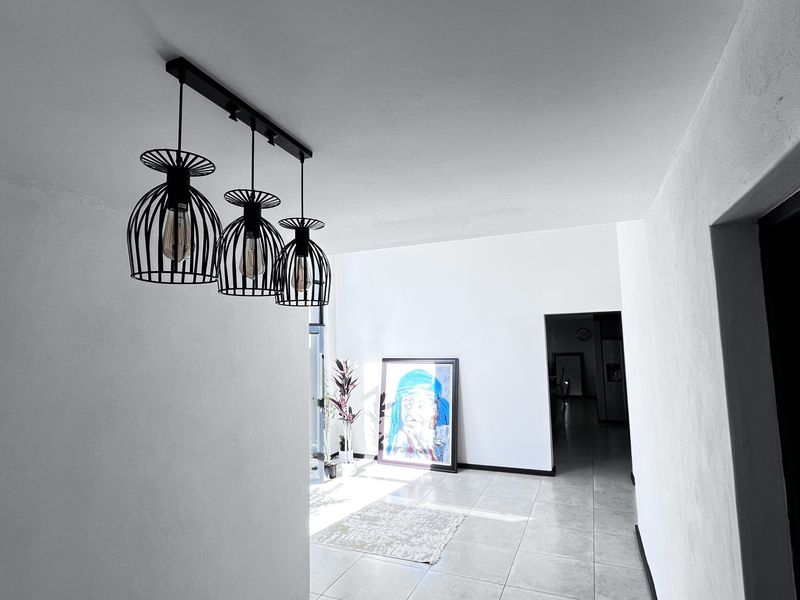 A bright, open-plan living area featuring modern industrial-style pendant lighting and neutral tiled flooring, leading towards an adjacent room.