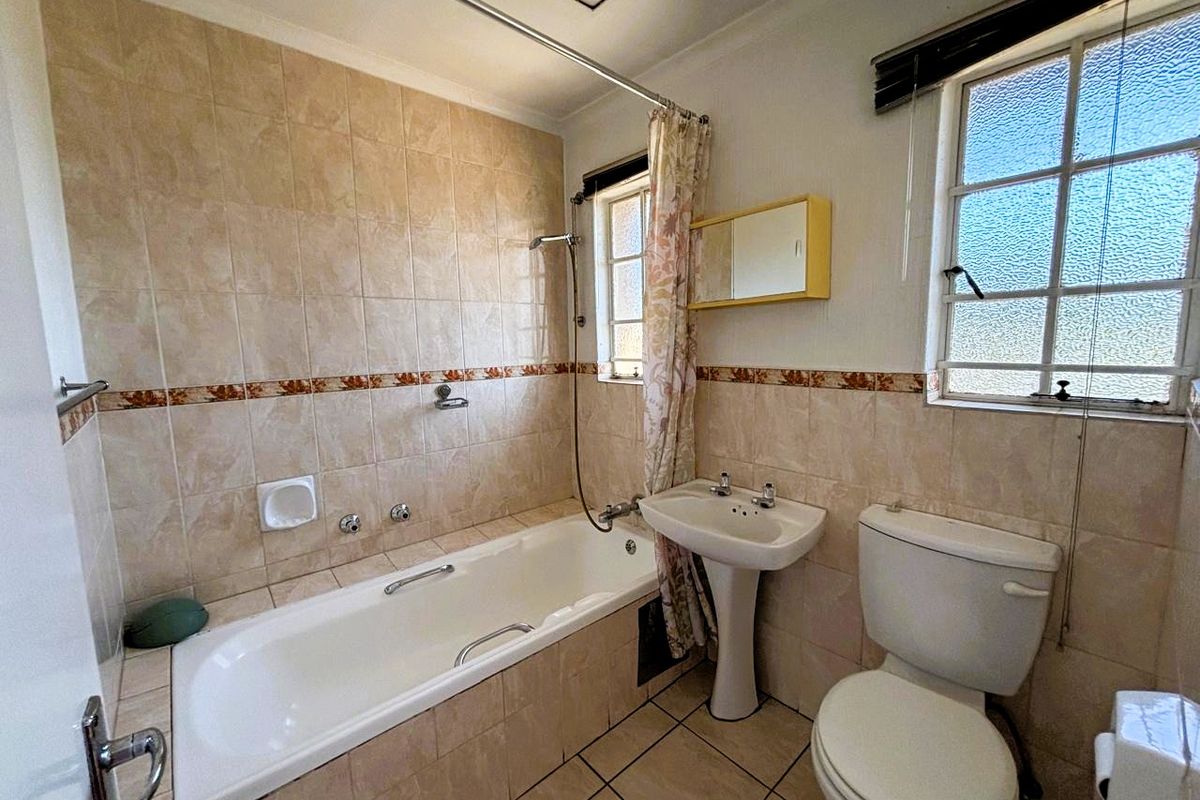 A functional bathroom featuring a bathtub with shower attachment, pedestal basin, and toilet, finished with neutral wall tiles and natural light from two windows.