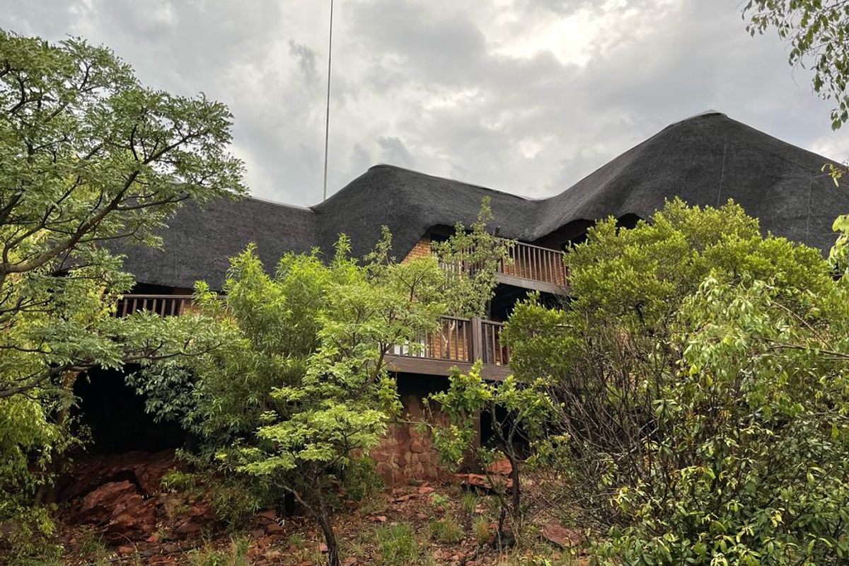 A multi-level house featuring a traditional thatched roof and wooden balconies, nestled within a natural, bushveld-style garden setting.